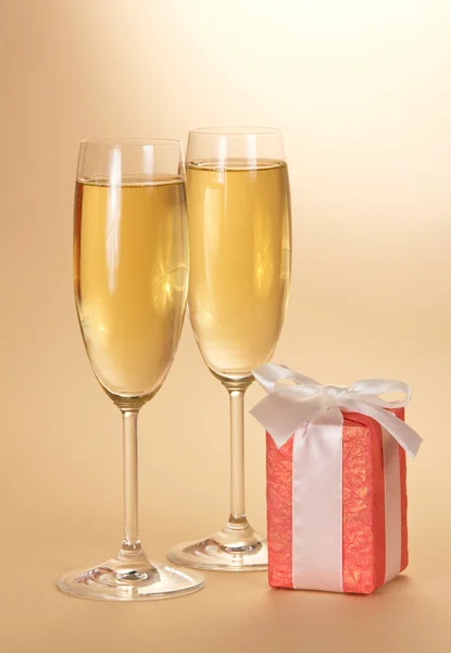 Two wine glasses with champagne and a gift box with a white bow on a beige background — Stock Photo, Image