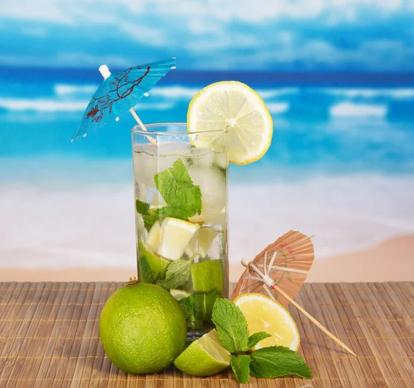 Das Cocktailglas, ist mit Sonnenschirmen und einer Zitrusfrucht auf einem Bambustuch gegen das Meer verziert — Stockfoto
