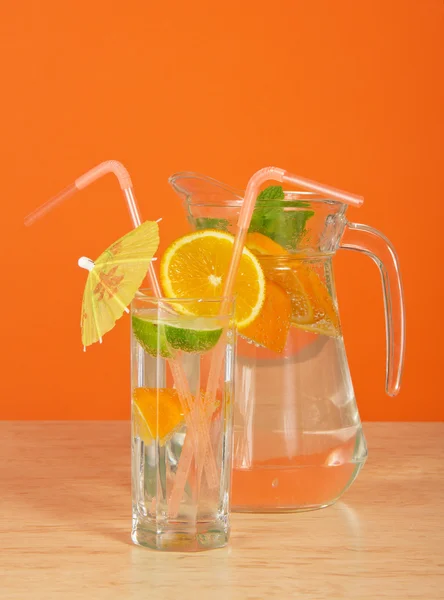 O copo de bebida com uma palha, decora-se com um guarda-chuva e um segmento cor-de-laranja, um jarro em uma mesa — Fotografia de Stock