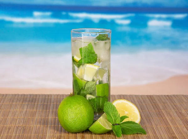 Glass with a mojito, a juicy lime and a spearmint leaf, on a bamboo cloth against the sea — Stock Photo, Image
