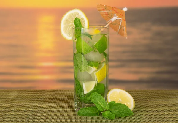 Cold drink with a lemon slice, spearmint, on a green bamboo cloth against the sunset — Stock Photo, Image