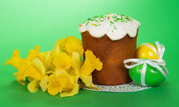 Bouquet of yellow narcissuses, Easter cake on a napkin and eggs with ribbons, on a green background — Stock Photo, Image