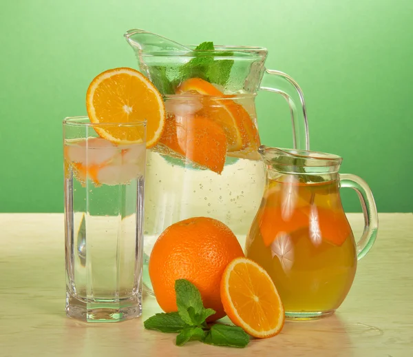 Jugs with drinks, a glass and oranges on a table — Stock Photo, Image