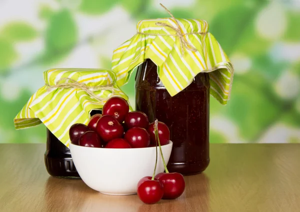 Two jars of jam, sweet cherries in a ceramic cup and two berries on a table — Stock Photo, Image