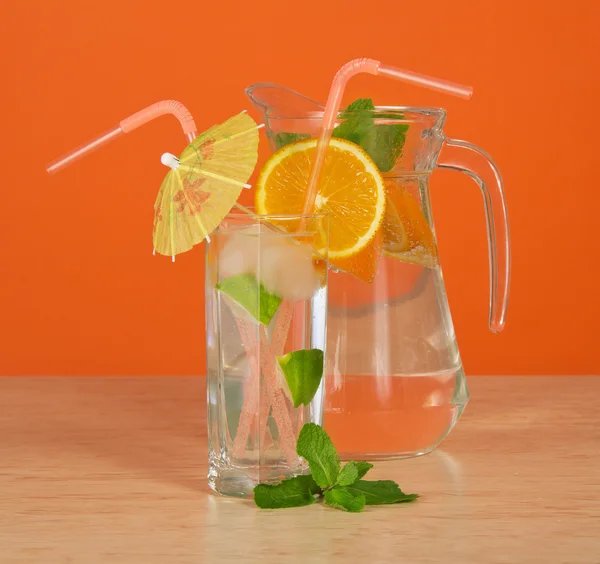 El vaso de bebida con una paja, está decorado con un paraguas y un segmento naranja, una jarra sobre una mesa —  Fotos de Stock