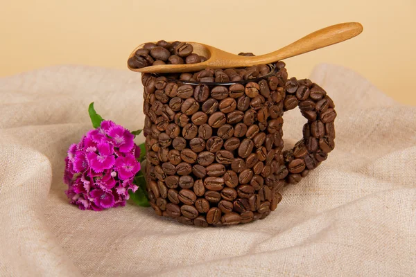 Wooden spoon, cup from coffee grains and a branch of bright carnation, on a sackcloth — Stock Photo, Image