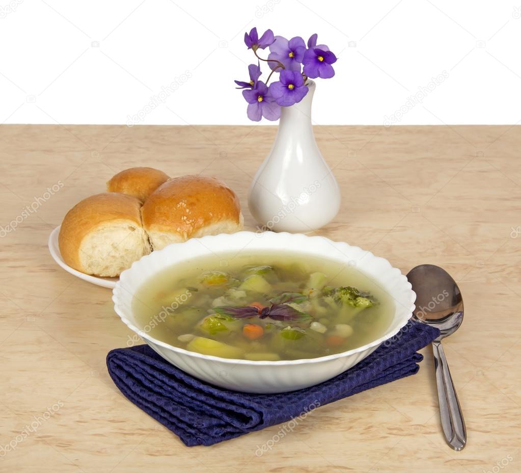 Vase with violets, a plate of vegetable soup, a spoon and a saucer with pies on a table