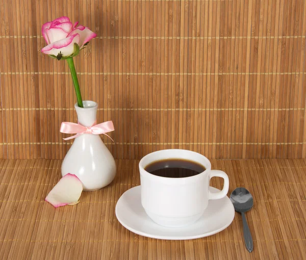 Vase with a rose, decorated with a bow, a cup with drink against a bamboo cloth — Stock Photo, Image