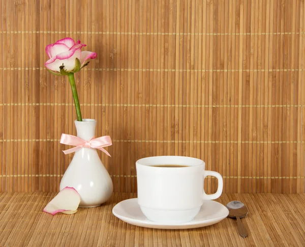 Vase with a rose, decorated with a bow, a cup with drink against a bamboo cloth — Stock Photo, Image