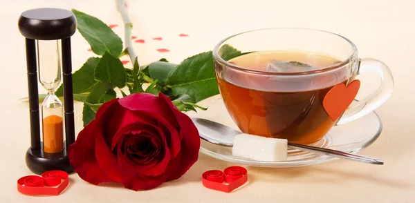 Горячий чай, красная роза и песочные часы на ткани, украшенной сердцами — стоковое фото