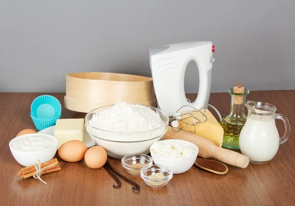 Cottage cheese, sour cream, cheese, butter, mixer and cake pan on a table, on a gray background — Stock Photo, Image