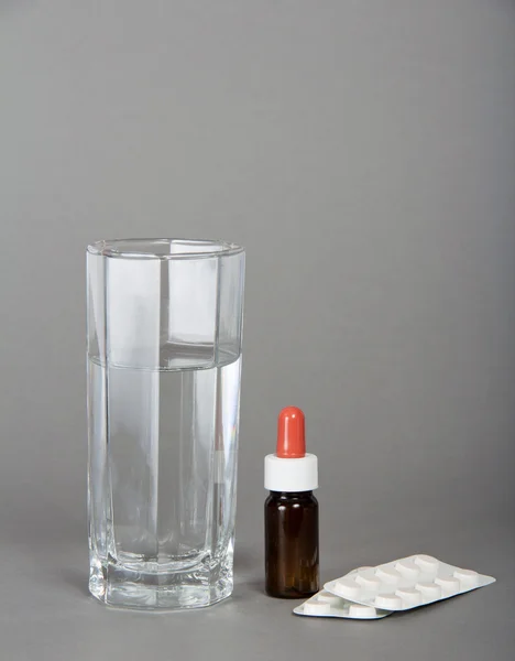 Kaca dengan air, tablet dan botol dengan tetes pada abu-abu — Stok Foto