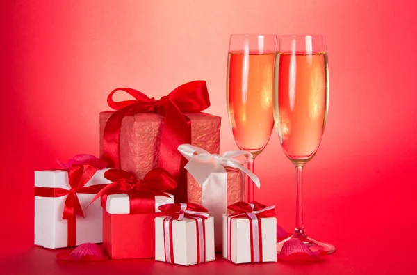 Two glasses of champagne, gift boxes with tapes and bows and petals of flowers on a red background — Stock Photo, Image