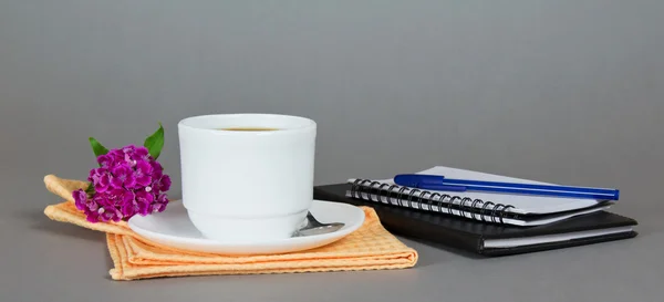 Kopje koffie met een schotel, een lepel en een bloem op een servet, een schetsblok en de bal-punt op een grijze achtergrond — Stockfoto