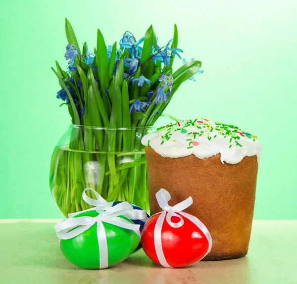 Easter cake, eggs with ribbons and a vase with the flowers on a table on a green background — Stock Photo, Image