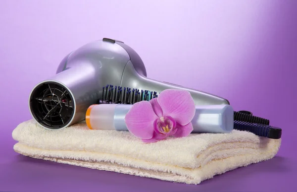 The hair dryer, hairbrush, shampoo on a towel, decorated with a flower, on the violet — Stock Photo, Image