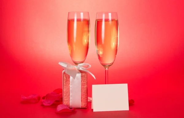 Two wine glasses with champagne, a gift box with a white bow, rose petals and blank card on a red background — Stock Photo, Image