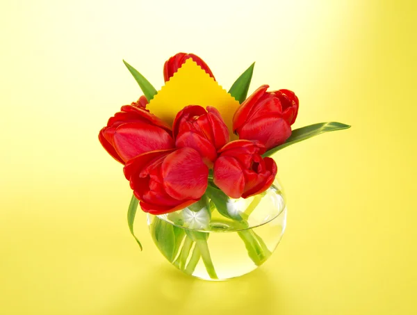 Red tulips in a round transparent vase and a card, on the yellow — Stock Photo, Image