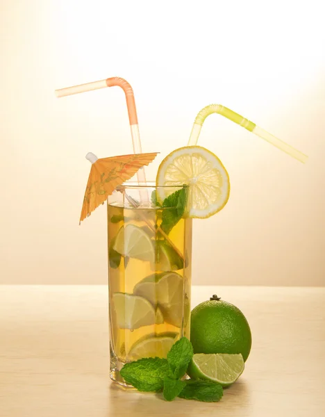 Beber con una pajita, está decorado con una rebanada de un limón y un paraguas, una lima madura y menta, sobre una mesa beige — Foto de Stock