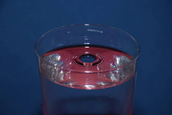 青い背景に飛び散った液滴と それに赤い反射を持つ水のガラスの閉鎖 — ストック写真