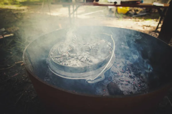 A Dutch Oven stands on hot, glowing coals in the smoke. Cooking on fire in the great outdoors.