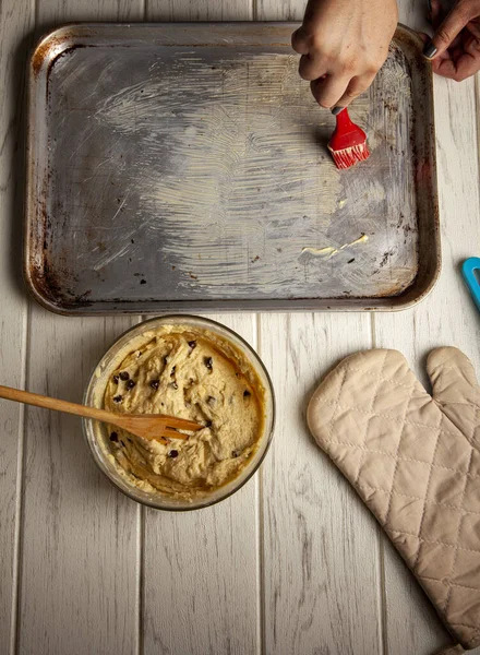 Overhead shot of spreading baking tray with butter, raw dough of chocolate chip cookies in a bowl with a wooden fork, oven glove on the wooden table