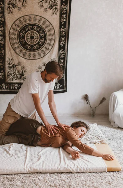 一名年轻的白种人胡子按摩师和一名妇女在家里的垫子上做泰式瑜伽按摩 — 图库照片