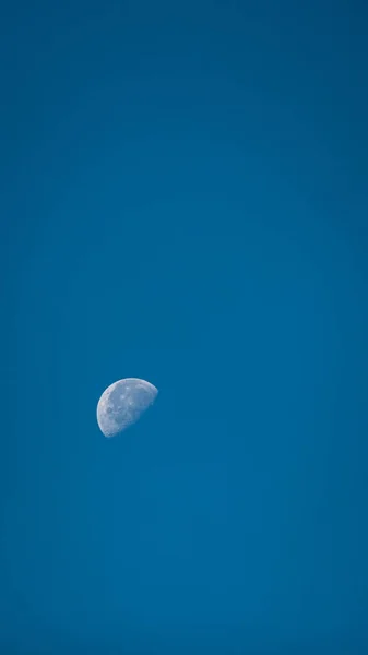 A vertical shot of the half moon in the blue sky.