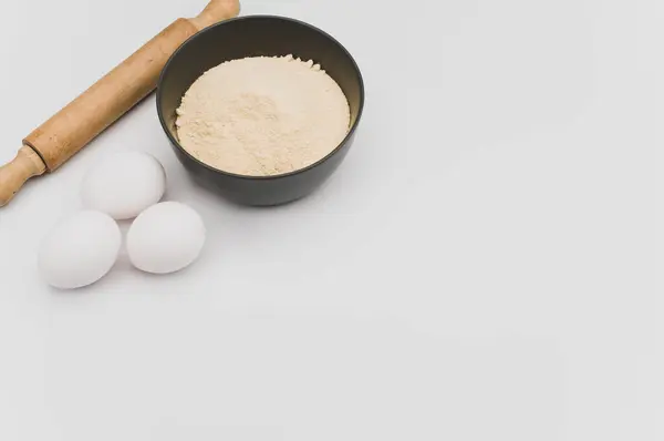 stock image wooden rolling pin white eggs and dark gray bowl with wholegrain flour on white background with copy space