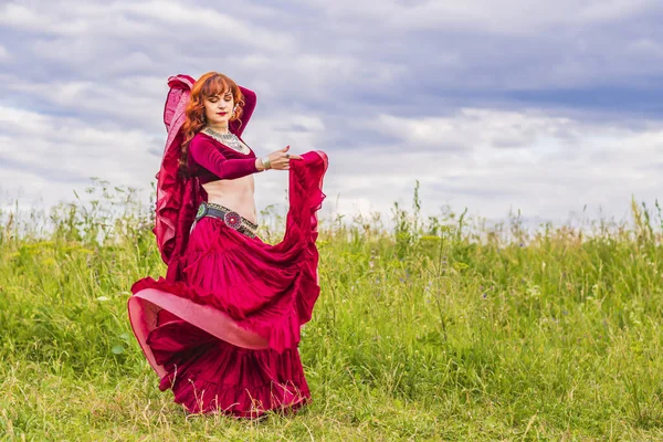 穿着红衣服的年轻漂亮的红头发姑娘在碧空荡荡的草地上欢快地跳着迷人的舞曲 — 图库照片