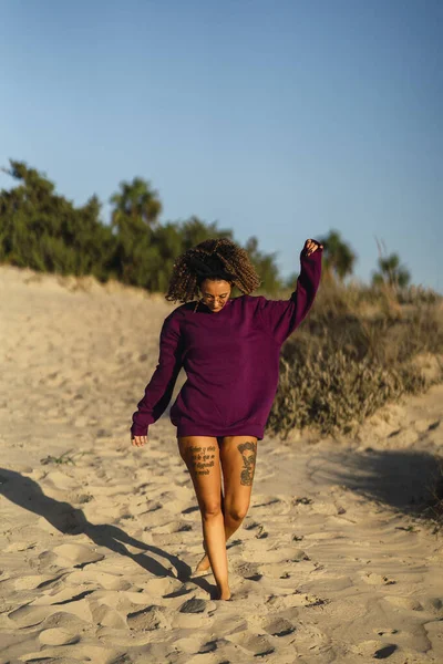 A vertical shot of a sexy tattooed female in a long purple sweater posing on a beach