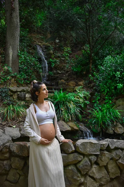 一位漂亮的年轻白人孕妇 身穿白衣 在大自然中摆姿势 靠着石墙 — 图库照片
