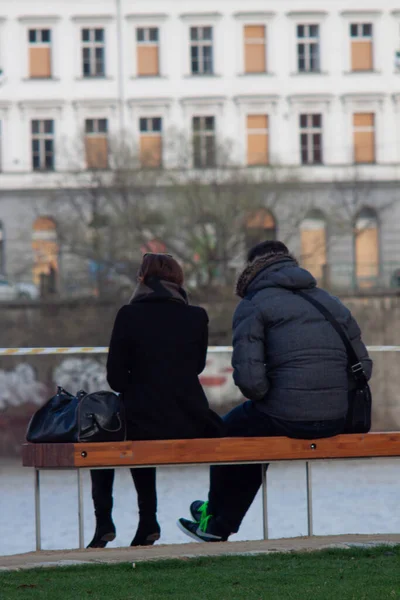 冬天有两个人坐在一条木制长椅上 前面是水 另一边是一座有许多窗户的建筑物 — 图库照片