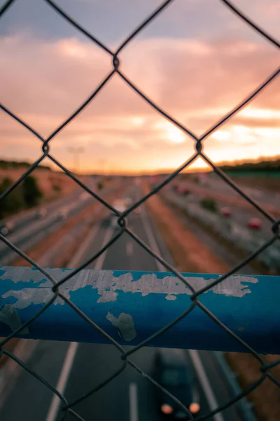 A vertical shot of a metallic fence on a background of a highway in the evening.