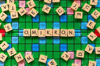 Scrabble tokens form the word omikron on the green original game board in middle of other single tokens, horizontal shot, German version clipart