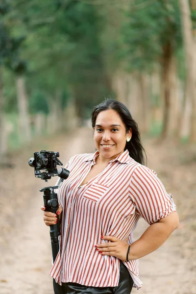 A smiling Hispanic woman holding a camera for a vlog against a blurred background