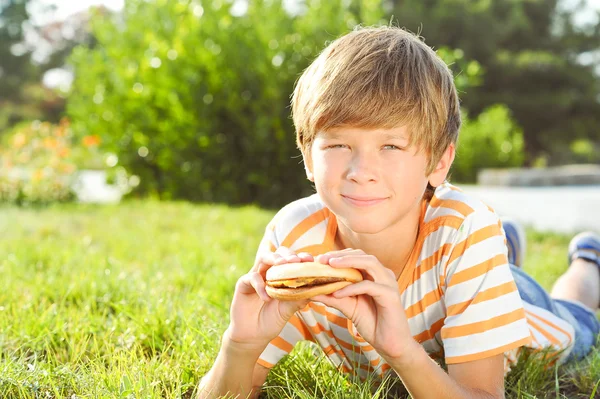 Criança deitada na grama comendo hambúrguer — Fotografia de Stock