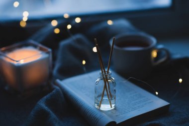 Liquid home freagrance in glass bottle with wooden sticks on paper book with burning candle and cup of tea on knit cloth close up. Winter cozy season. Aromatherapy. 