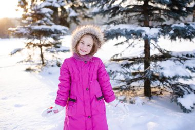 Smiling kid girl 5-6 year old wear pink winter jacket in snowy park outdoors. Looking at camera. Childhood. 