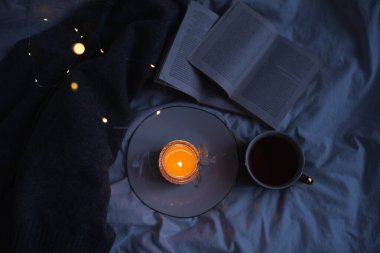 Burning scented candle on ceramic tray with open paper book and knit sweater cloth in bed. Winter cozy hygge atmosphere with cup of black coffee at home. Coziness. 