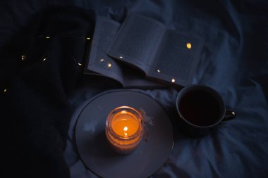 Burning scented candle on ceramic tray with open paper book and knit sweater cloth in bed. Winter cozy hygge atmosphere with cup of black coffee at home. Coziness. 