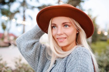 Beautiful blonde smiling adult girl 20-24 year old wear casual warm clothes and hat posing in city park. Autumn season. Romantic lady outdoors.  