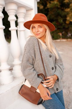 Stylish blonde young adult girl 22-24 year old wear felt hat, denim pants and knit woolen cardigan holding trendy leather bag walk in city street posing over sunset background outdoors close up. 20s.