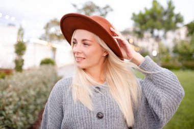 Smiling young beautiful blonde woman wear hat and knitted woolen jumper posing over city street in park outdoors close up. Happy pretty teenage girl 18-19 year old having fun outside. 20s. Happiness. 