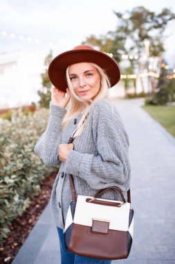 Happy stylish blonde girl 19-20 year old wear hat, knit woolen jumper and holding leather bag walk in park outdoors. Looking at camera. Happiness. Autumn season. 