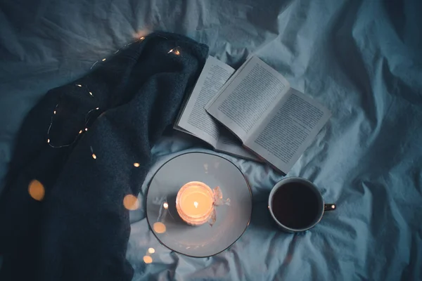 Burning scented candle on ceramic tray with open paper book and knit sweater cloth in bed. Winter cozy hygge atmosphere with cup of black coffee at home. Coziness. 