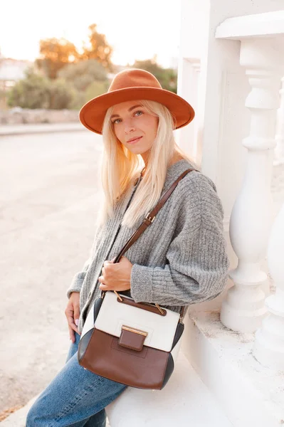 Stylish blonde young adult girl 22-24 year old wear felt hat, denim pants and knit woolen cardigan holding trendy leather bag walk in city street posing over sunset background outdoors close up. 20s.