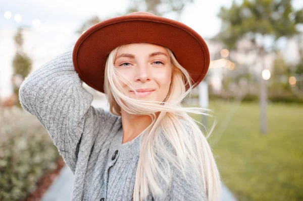 Beautiful blonde smiling adult girl 20-24 year old wear casual warm clothes and hat posing in city park. Autumn season. Romantic lady outdoors. Looking at camera. 