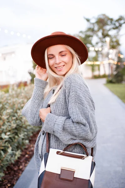 Laughing blonde cute young girl 20-24 year old wear hat, knitted woolen gray sweater and holding trendy leather bag walk in park outdoors over urban city street background. Positive emotions. 