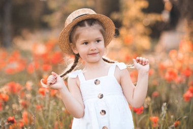 3-4 yaşlarında gülümseyen bir bebek hasır şapka ve beyaz kırsal bir elbise giyer ve doğal afyon arkaplanı üzerinde poz verir. Çiçek çayırındaki mutlu çocuk. Bahar geldi. Çocukluk. 